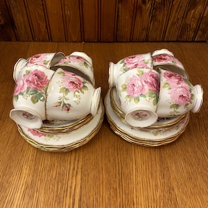 Puede incluir: Un juego de seis tazas de té y platillos con un patrón floral con rosas rosadas. Las tazas y los platillos tienen un borde dorado.