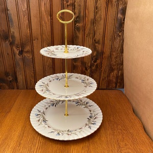 May include: Three-tiered cake stand with a gold metal frame and white porcelain plates. Each plate features a floral pattern with blue and green accents.