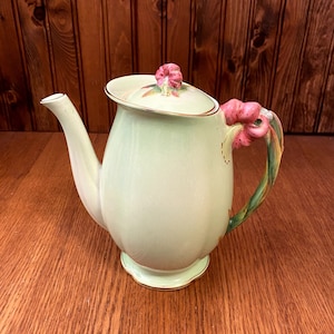 May include: A light green ceramic teapot with a floral design. The teapot has a lid with a pink flower on top and a handle shaped like a pink flower with green leaves.