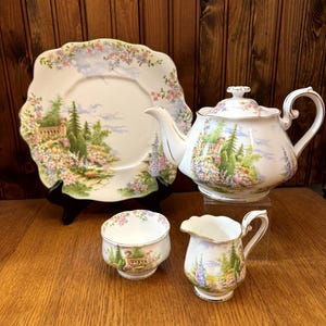 Pode incluir: Um conjunto de chá de porcelana vintage com bule, cremeira, açucareiro e prato. O conjunto tem um design floral e paisagístico com cores pastel. O prato é recortado e o bule tem uma alça curva.