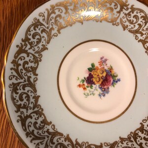 May include: A light blue saucer with a gold floral design around the edge. The center of the saucer has a white circle with a floral design in the center.