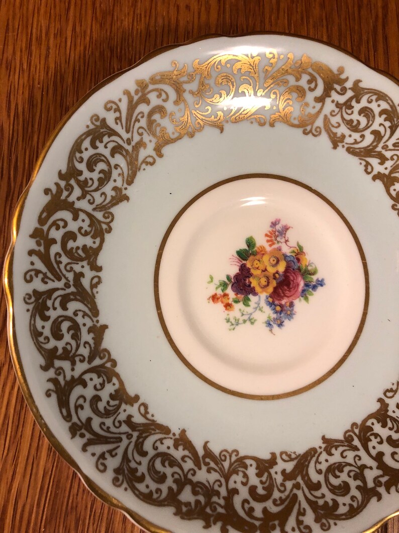 May include: A light blue saucer with a gold floral design around the edge. The center of the saucer has a white circle with a floral design in the center.