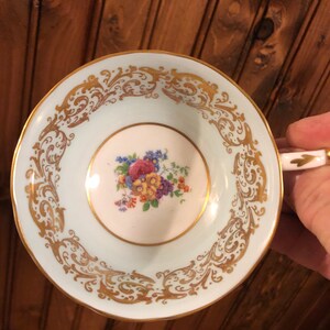 May include: A light blue teacup with a gold floral design around the rim and a floral pattern in the center. The cup is made of porcelain and has a delicate, feminine feel.