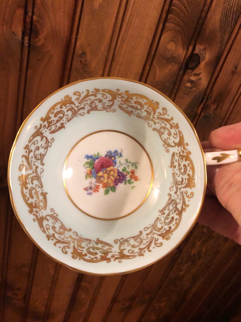 May include: A light blue teacup with a gold floral design around the rim and a floral pattern in the center. The cup is made of porcelain and has a delicate, feminine feel.