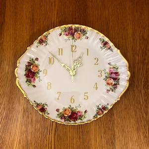 Puede incluir: Un reloj decorativo de plato blanco con manecillas y números dorados. El plato tiene un borde festoneado con ribete dorado y está decorado con motivos florales en tonos rojos, rosas y amarillos. El reloj está sobre un fondo de madera.