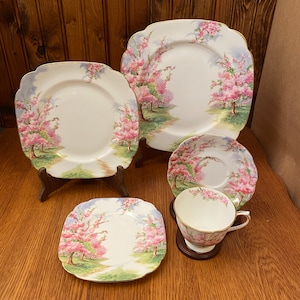May include: A set of square-shaped porcelain plates with a floral design featuring pink blossoms and green leaves. The plates are white with a gold trim. The set includes a teacup and saucer.