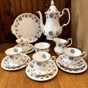 Puede incluir: Un juego de té de porcelana blanca con un estampado floral en morado y dorado. El juego incluye una tetera, una jarra de crema, un azucarero, una fuente y varias tazas de té con platillos. El juego de té está dispuesto sobre una superficie de madera.