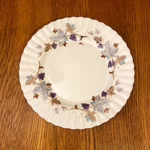 May include: A white ceramic dinner plate with a scalloped edge and a grapevine design in shades of purple, blue, and brown.