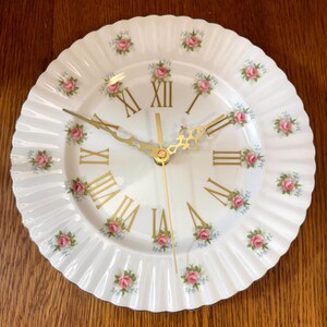 May include: White ceramic wall clock with a ruffled edge and gold hands. The clock face features gold Roman numerals and pink rose floral accents. A decorative piece for the home.