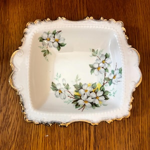 May include: A square, white porcelain dish with a floral design. The dish has a gold trim and a pattern of white flowers with yellow centres and green leaves. The dish is set against a wooden surface.