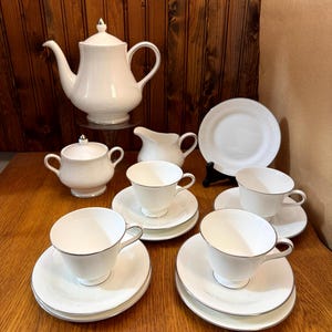 Könnte beinhalten: Ein weißes Teeservice mit Teekanne, Milchkännchen, Zuckerdose und vier Tassen und Untertassen. Das Set hat ein zartes Blumenmuster und einen silbernen Rand.