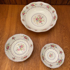 May include: Three white ceramic bowls with a floral pattern. The bowls are nested inside each other, with the largest bowl on the bottom and the smallest bowl on top. The bowls have a scalloped edge and a gold trim.