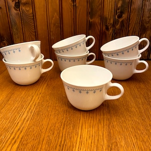 May include: Six white ceramic mugs with blue snowflake designs. The mugs are stacked in pairs and are arranged on a wooden surface.