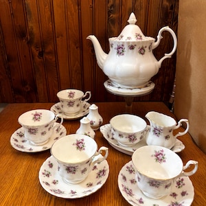 Puede incluir: Un juego de té de porcelana blanca con diseños florales morados. El juego incluye una tetera, tazas, platillos, una crema y saleros y pimenteros. El juego de té se exhibe sobre una superficie de madera.