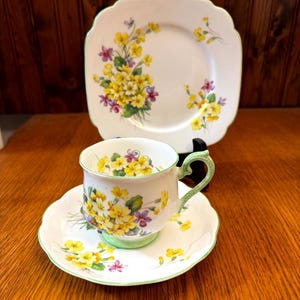 May include: A vintage teacup, saucer, and square plate set. The white porcelain pieces feature a floral design with yellow and purple flowers, accented with green trim. The teacup has a curved handle.