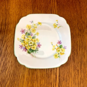 May include: A white square plate with a green rim features a floral design of yellow, purple, and green primroses.