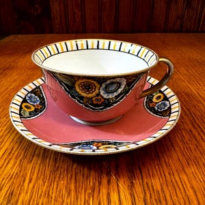 May include: A pink teacup and saucer set. The cup and saucer have a floral design with yellow, blue, and white flowers. The rim of the cup and saucer has a black, yellow, and white striped pattern. The handle of the cup is gold.