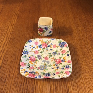 May include: A white square dish with a floral pattern in shades of pink, blue, yellow, and green. A smaller square dish with a similar floral pattern is placed on top of the larger dish.