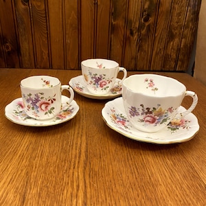 Peut inclure: Trois tasses à thé et soucoupes fleuries avec une bordure dorée. Les tasses et les soucoupes ont un fond blanc avec un motif floral coloré.