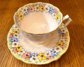 May include: A vintage teacup and saucer set. The cup and saucer are decorated with a floral pattern in yellow, blue, pink, and white. The edges of the cup and saucer are scalloped and trimmed with gold. The set is placed on a wooden surface.