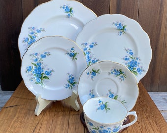 May include: A set of white porcelain teacups and saucers with a delicate blue forget-me-not floral pattern. The set includes a teacup, saucer, and two larger plates.