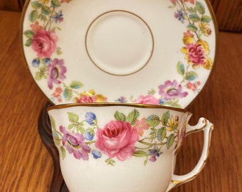 May include: A white teacup and saucer with a gold rim and floral pattern. The pattern features pink, purple, and yellow flowers with green leaves.