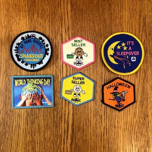 Puede incluir: Una colección de seis parches coloridos con varios diseños y texto. Los parches incluyen un parche azul y blanco "Girl Guides Shakeout Challenge", un parche rosa y blanco "Mint Seller" y un parche azul y amarillo "It's a Sleepover".