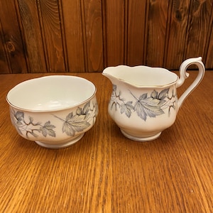 May include: A white sugar bowl and creamer set with a gray and green leaf design. The set is made of fine china and has a gold trim.
