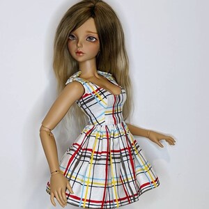 May include: A doll wearing a white dress with a red, yellow, and blue plaid pattern. The dress has a sweetheart neckline and a fitted bodice. The doll has long, blonde hair.