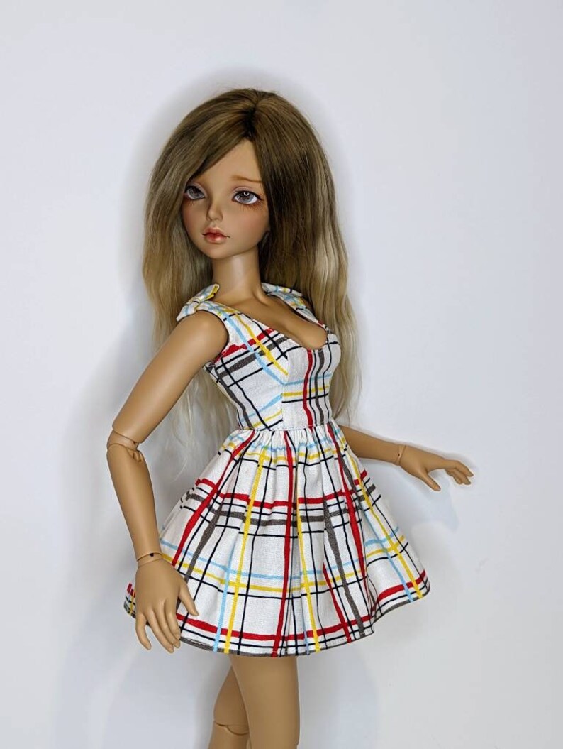 May include: A doll wearing a white dress with a red, yellow, and blue plaid pattern. The dress has a sweetheart neckline and a fitted bodice. The doll has long, blonde hair.