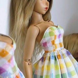 May include: A doll wearing a white dress with a colorful plaid pattern. The dress has a sweetheart neckline and a gathered skirt. The doll has long blonde hair and is looking to the side.