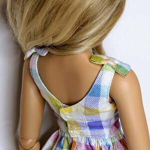 May include: A doll wearing a sleeveless dress with a plaid pattern in shades of blue, green, yellow, and purple. The dress has a white background and a glittery texture. The doll has long blonde hair.