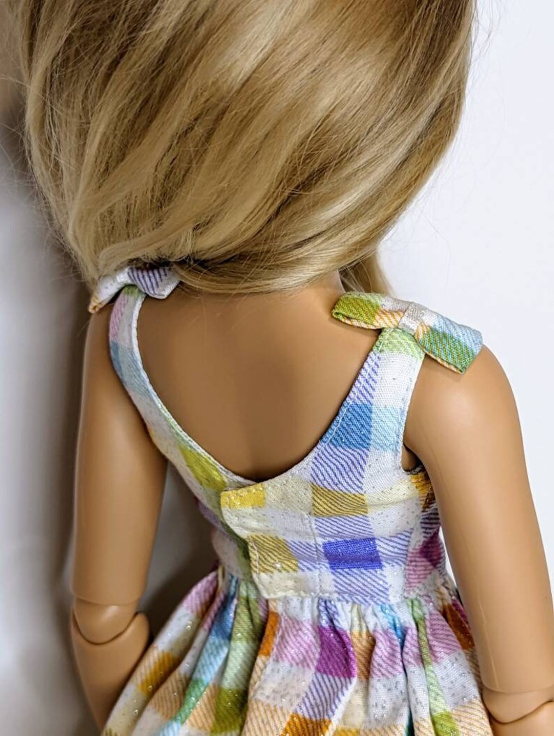 May include: A doll wearing a sleeveless dress with a plaid pattern in shades of blue, green, yellow, and purple. The dress has a white background and a glittery texture. The doll has long blonde hair.