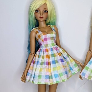 May include: A doll wearing a white dress with a colorful plaid pattern. The dress has a sweetheart neckline and a full skirt. The doll has long, wavy, yellow and green hair.