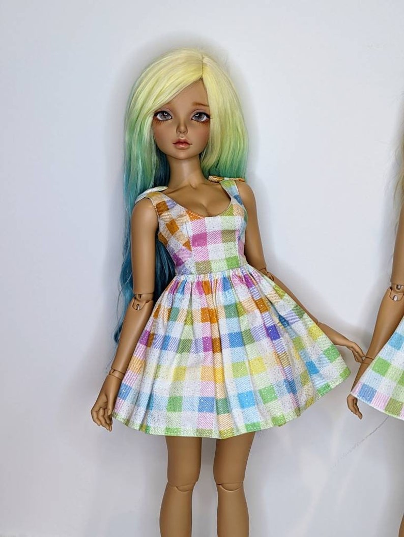 May include: A doll wearing a white dress with a colorful plaid pattern. The dress has a sweetheart neckline and a full skirt. The doll has long, wavy, yellow and green hair.