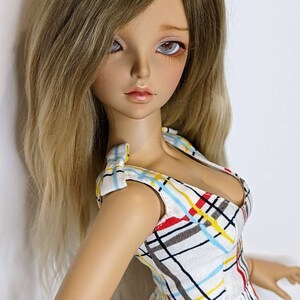 May include: A doll with long, wavy, light brown hair wearing a white dress with a plaid pattern in red, yellow, and blue.