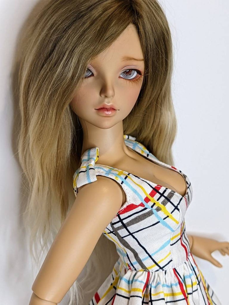 May include: A doll with long, wavy, light brown hair wearing a white dress with a plaid pattern in red, yellow, and blue.