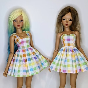 May include: Two dolls wearing colorful gingham dresses. The dresses have a white background with squares of pink, blue, yellow, and orange. The dolls have long hair and are standing on a white background.