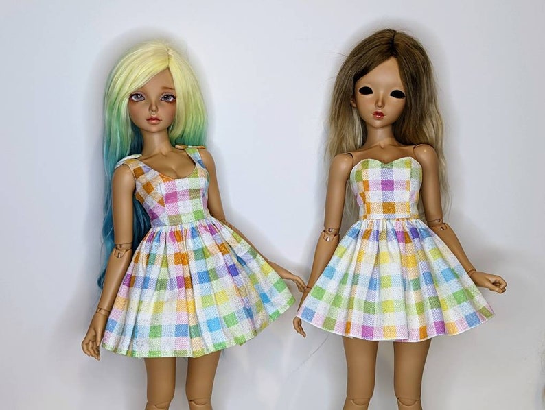 May include: Two dolls wearing colorful gingham dresses. The dresses have a white background with squares of pink, blue, yellow, and orange. The dolls have long hair and are standing on a white background.