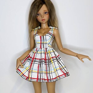 May include: A doll wearing a white, red, yellow, and blue plaid dress with a sweetheart neckline and bow straps.