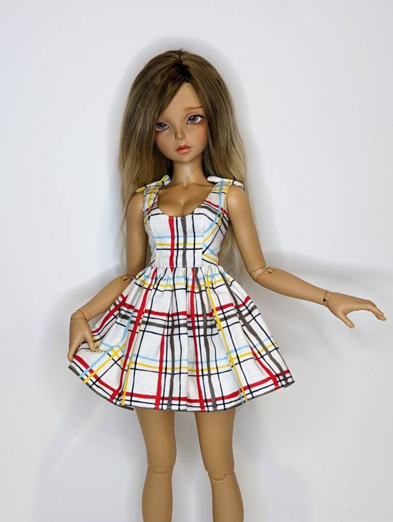 May include: A doll wearing a white, red, yellow, and blue plaid dress with a sweetheart neckline and bow straps.
