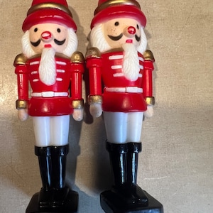 May include: Two red and white plastic nutcracker ornaments with gold accents. The nutcrackers are wearing black boots and have white beards and mustaches.