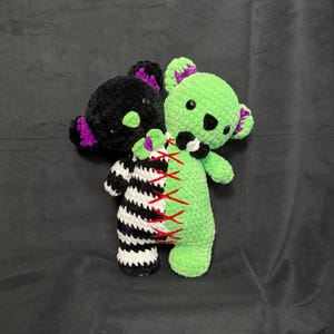 May include: Two crocheted teddy bears sewn together. One bear is black and white striped, the other is green. The bears are stitched together with red thread.