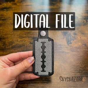 May include: A black and gray key chain shaped like a razor blade. The keychain has a silver ring for attaching keys.