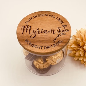 Könnte beinhalten: Ein klares Glas mit einem Holzdeckel. Auf dem Deckel steht der Name "Myriam" und die Worte "Gute Besserung, Liebe" und "Wünscht Dir Ursel". Das Glas enthält goldverpackte Pralinen.