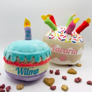 Könnte beinhalten: Zwei Plüsch-Geburtstagskuchen-Spielzeuge mit weichen Texturen. Der linke Kuchen ist hellblau mit einer lila Kerze, die die Zahl 2 und den Namen Wilma zeigt. Der rechte Kuchen ist weiß mit bunten Streuseln, roten und grünen Kerzen und dem Namen Vavaria.