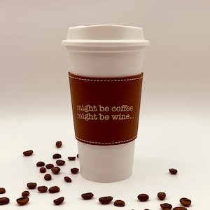 Könnte beinhalten: Weißer To-Go-Becher mit einer braunen Lederhülle, auf der in goldenen Buchstaben "might be coffee might be wine..." steht. Der Becher ist von Kaffeebohnen umgeben.