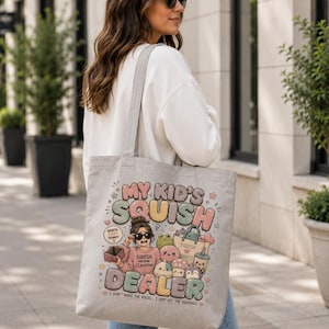 May include: A light gray canvas tote bag featuring a colorful graphic print with the text "MY KID'S SQUISH DEALER" and "I DON'T MAKE THE RULES... I JUST GET THE SQUISHES!" along with illustrations of a person and various small plush toys.