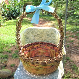 May include: A woven wicker basket with a light brown, orange, and yellow color scheme. The basket has a large, rounded handle with a light blue bow tied at the top.