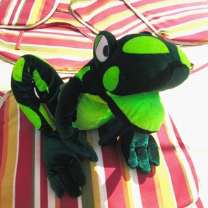 Pu&ograve; includere: Un giocattolo di peluche di rana verde e nero con grandi occhi rotondi. La rana &egrave; seduta su un cuscino a strisce rosso, giallo e bianco.
